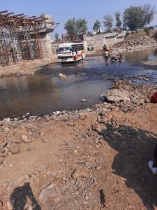 पानी और पत्थरों के बीच से निकलती बस और दुपहिया वाहन, भीमगढ़ नदी पर अधूरे पुल के कारण जोखिमभरा मार्ग।